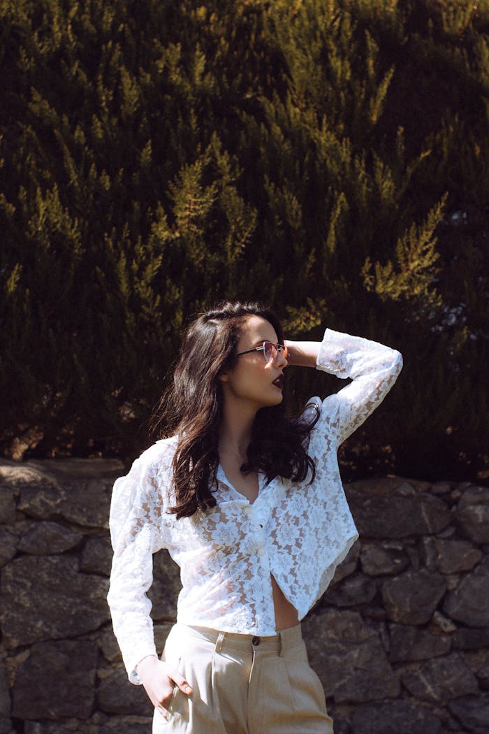 Fashionable woman in a white lace blouse posing confidently outdoors with greenery background.